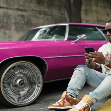 C. City sits on the ground, leaning against a classic pink car with large wire-spoke rims, looking at his phone.