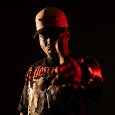 Casablanca the Gawd in low red light, pointing toward the camera, wearing a graphic t-shirt and a baseball cap.