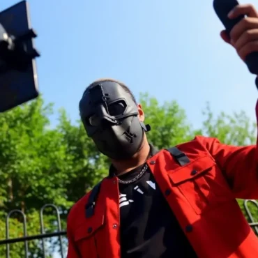 A person in a black hoodie and black face mask, partially lit by a red light against a dark background.