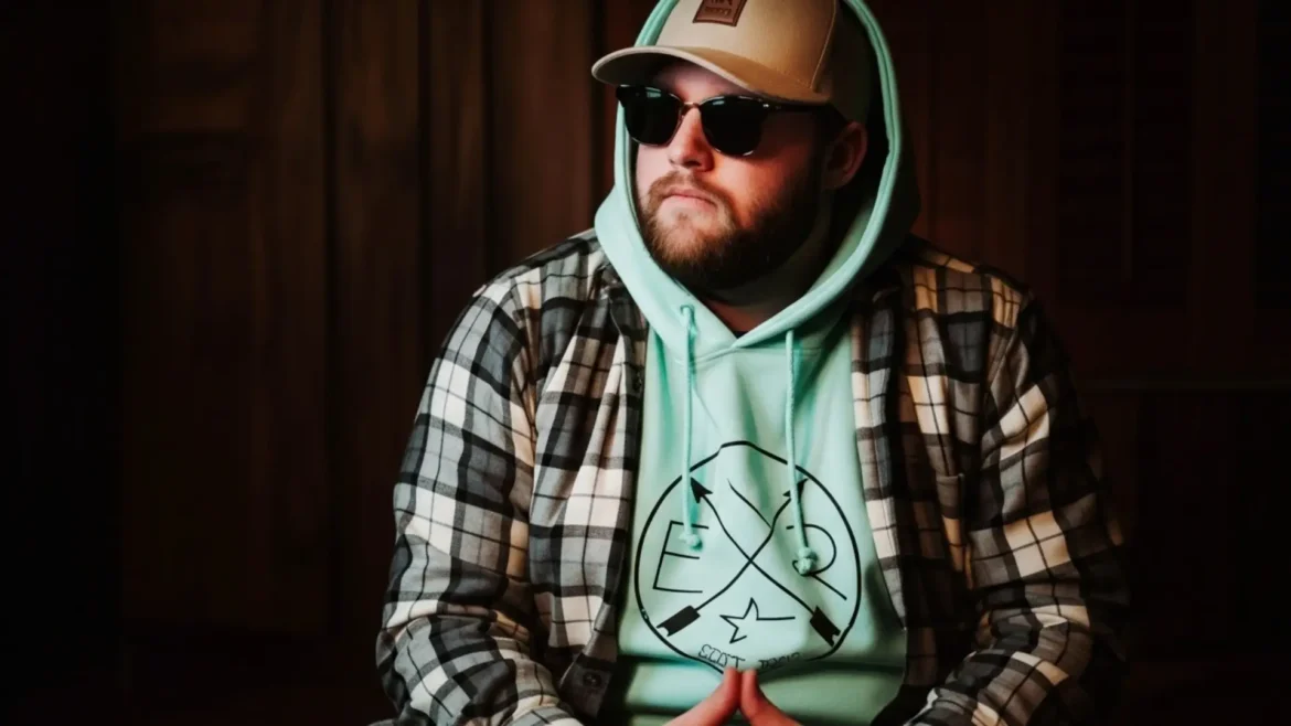Mr. Bronco, a rapper, sits indoors wearing a hat, sunglasses, and a plaid shirt over a hoodie, looking off to the side.