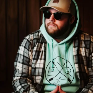 Mr. Bronco, a rapper, sits indoors wearing a hat, sunglasses, and a plaid shirt over a hoodie, looking off to the side.
