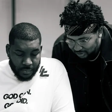A black and white photo of two men, likely Oryan Latin and his brother JuiceDaCity, looking down at a smartphone together in a studio setting.