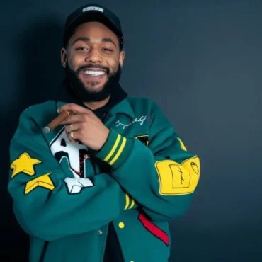 The Frost General, a Black man with a beard, wearing a green letterman jacket and a black cap, smiles at the camera while holding a cigar.