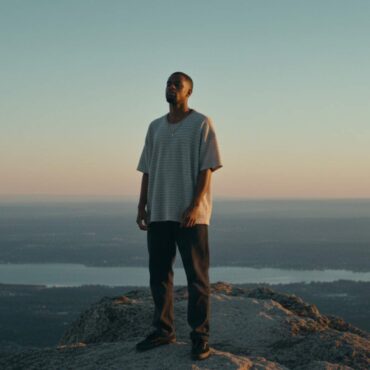 Kiran The Nomad standing on a mountain peak at sunset, looking over a valley.