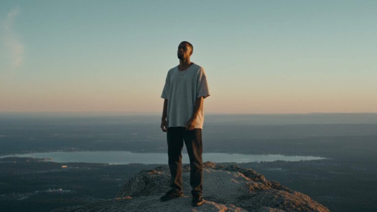 Kiran The Nomad standing on a mountain peak at sunset, looking over a valley.