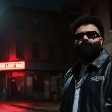 Sam Kays standing on a dark, wet city street at night, wearing a varsity jacket and sunglasses.