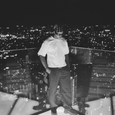 Black and white photo of rapper SIVH standing on a high-rise balcony at night with city lights in the background.
