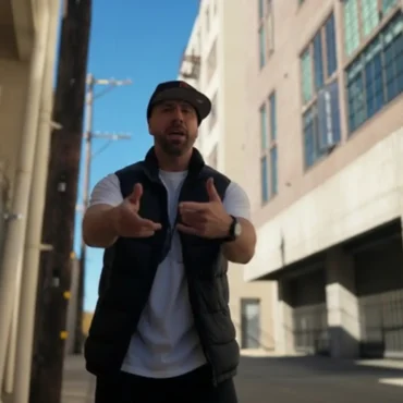 Tacer stands in an urban alleyway gesturing towards the camera, wearing a vest and a backward cap.