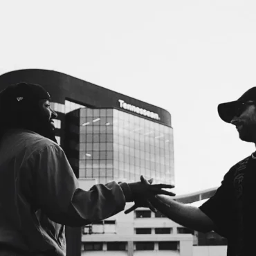 Two men, seen from a low angle, shaking hands outdoors with a modern glass building (The Tennessean) in the background.