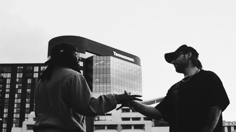 Two men, seen from a low angle, shaking hands outdoors with a modern glass building (The Tennessean) in the background.