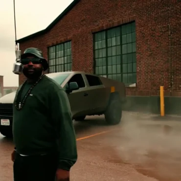 WizeTheChosen standing in front of a Cybertruck and a vintage microphone, wearing a bucket hat and sunglasses.