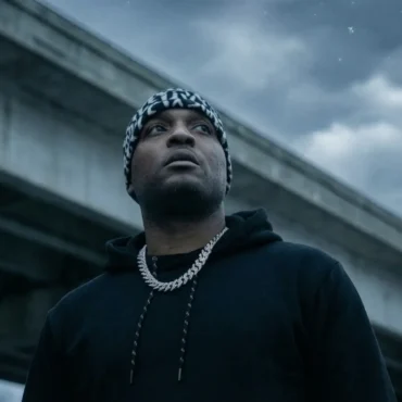 40 Glocc wearing a black hoodie and patterned beanie looking up while standing under a concrete overpass on a cloudy day.