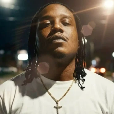 J.P with dreadlocks wearing a white t-shirt and gold cross chain standing in a street at night.