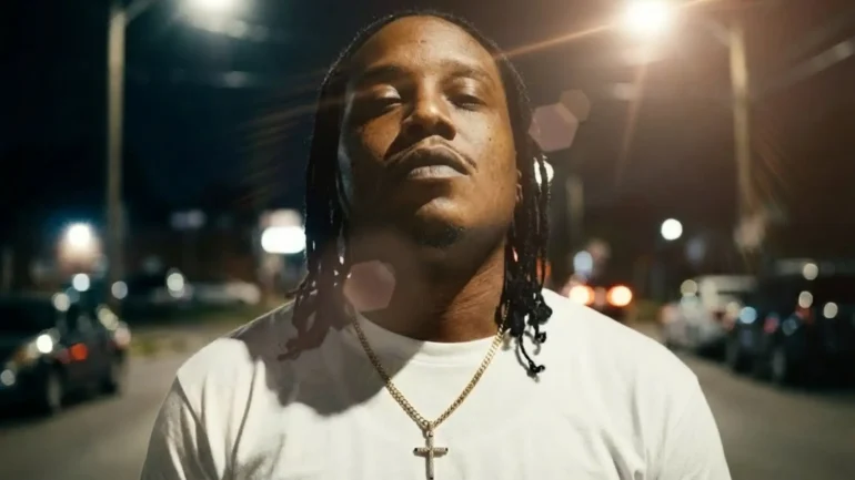 J.P with dreadlocks wearing a white t-shirt and gold cross chain standing in a street at night.