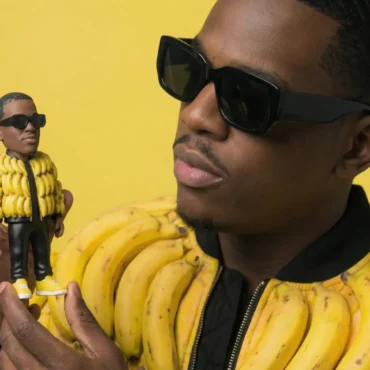 Black hip hop artist wearing sunglasses and a banana-peel bomber jacket holding a custom 3D vinyl toy of himself against a yellow background.