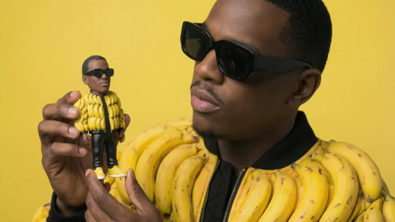 Black hip hop artist wearing sunglasses and a banana-peel bomber jacket holding a custom 3D vinyl toy of himself against a yellow background.