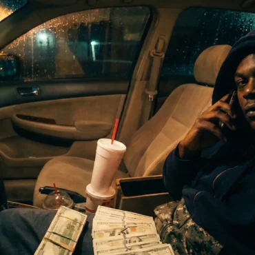 Amg Bubba sitting in a car counting a stack of cash holding a phone.