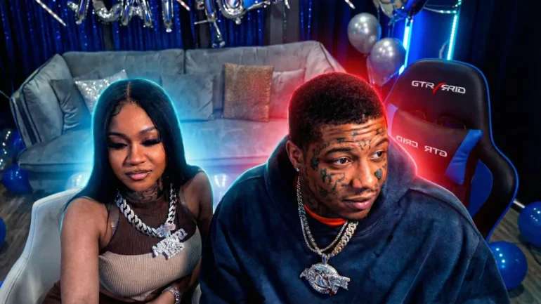 Blueface and Stunna Girl sitting closely together on a gray couch in a vibrant blue-lit gaming room decorated with balloons and streamers. Blueface, heavily tattooed with face ink, wears a dark hoodie and large gold chains, looking sideways with a serious expression. Stunna Girl, with long black hair and heavy makeup, sits beside him in a brown top and chunky "MONEY" necklace, smiling subtly. Neon blue and red lighting adds dramatic glow.