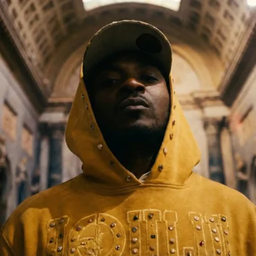 Cellz Grammz wearing a yellow studded hoodie standing in a building with high arches.