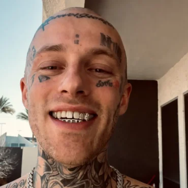 Rapper Free Zoey shirtless with face tattoos and silver grills smiling on a sunny balcony with palm trees in the background.