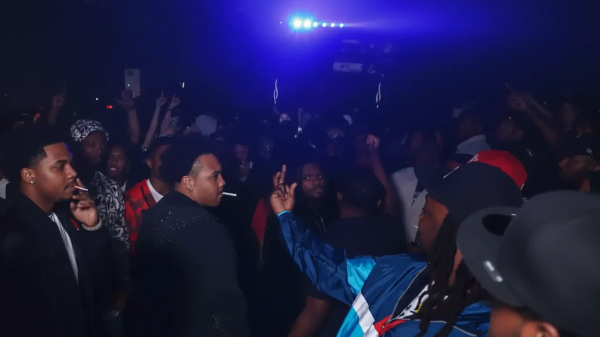 Blurry nightclub crowd under blue lights with a man in blue jacket throwing up a rake gang sign close to another man's face, capturing tense disrespect moment.