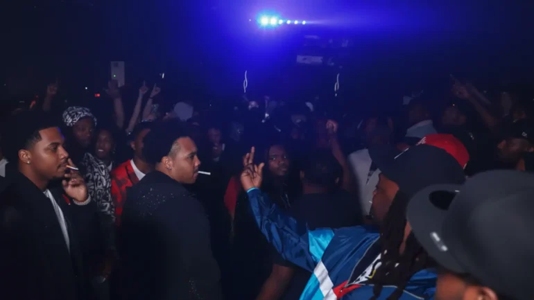 Blurry nightclub crowd under blue lights with a man in blue jacket throwing up a rake gang sign close to another man's face, capturing tense disrespect moment.