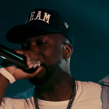 King Rizzy performing live on stage with a microphone wearing a black hat and gold chain.