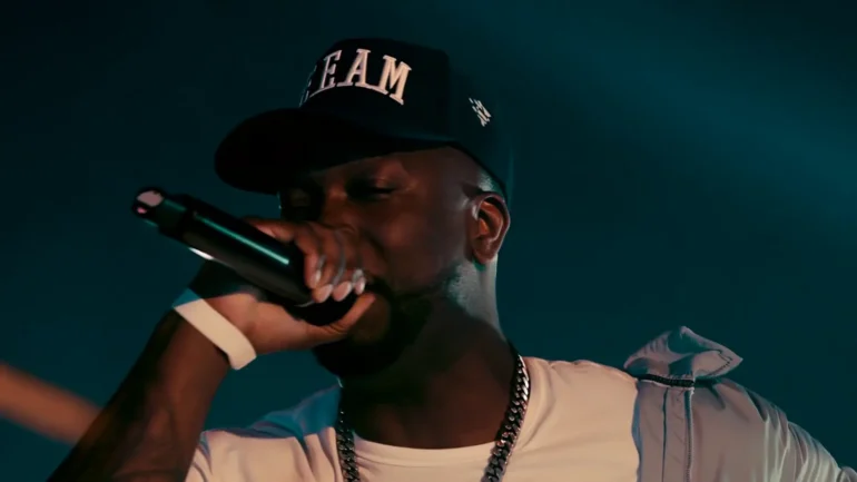 King Rizzy performing live on stage with a microphone wearing a black hat and gold chain.