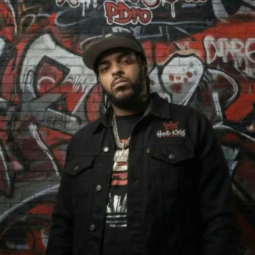 Rapper P~dro standing in front of a brick wall with Hood King Entertainment graffiti in red and black.