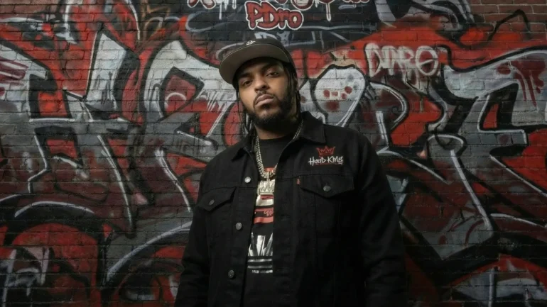 Rapper P~dro standing in front of a brick wall with Hood King Entertainment graffiti in red and black.