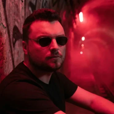 Ross Palmer wearing sunglasses in red neon lighting.