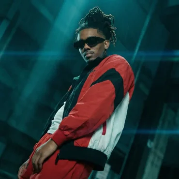 Trino rapper wearing red track jacket and sunglasses in teal light.