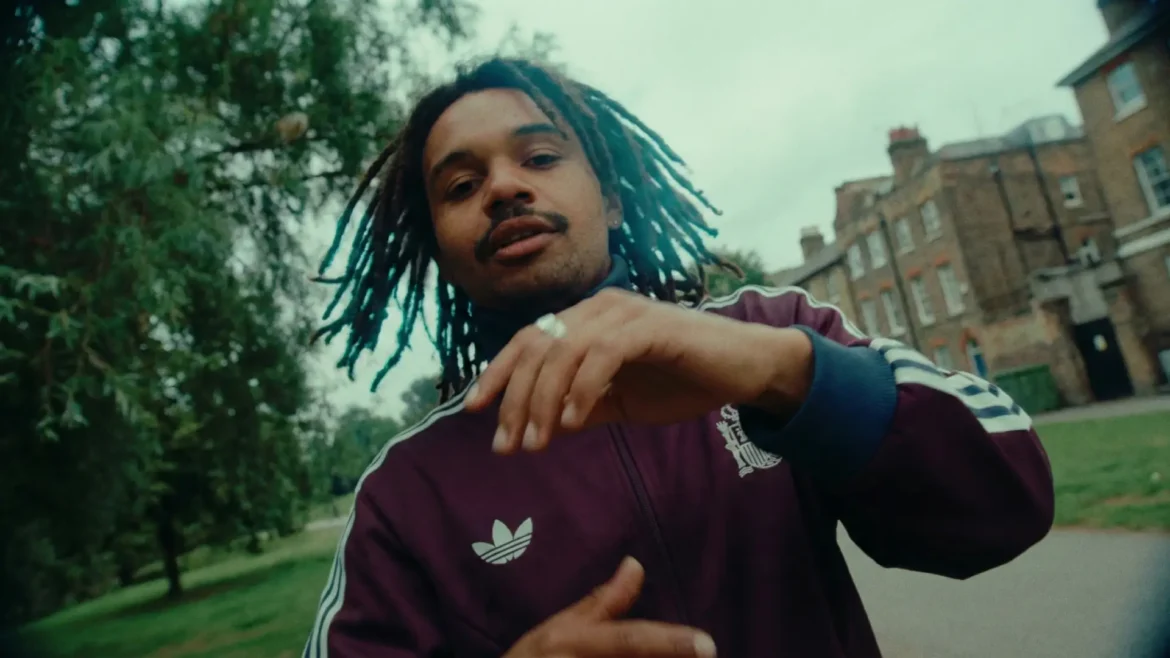 Rapper Yorke in a burgundy Adidas track jacket performing in an East London park with a fisheye lens effect.