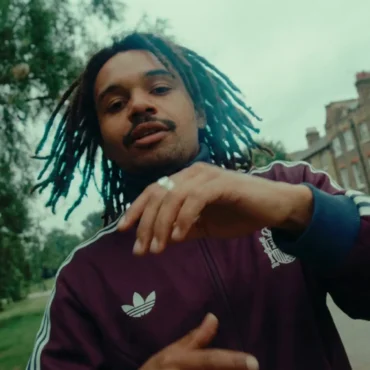 Rapper Yorke in a burgundy Adidas track jacket performing in an East London park with a fisheye lens effect.