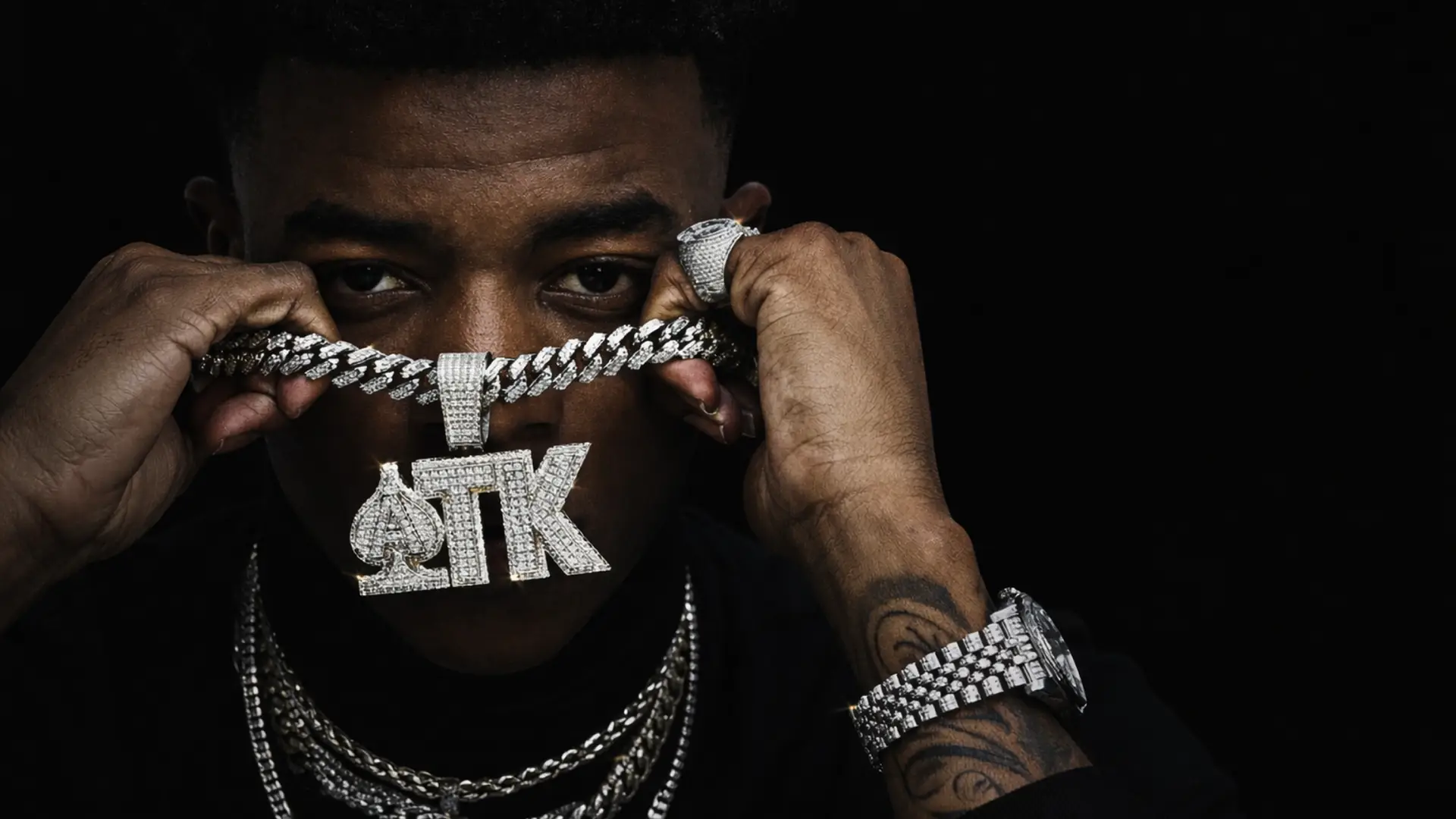 Rapper Yungeen Ace holding diamond ATK chain over face, intense stare.