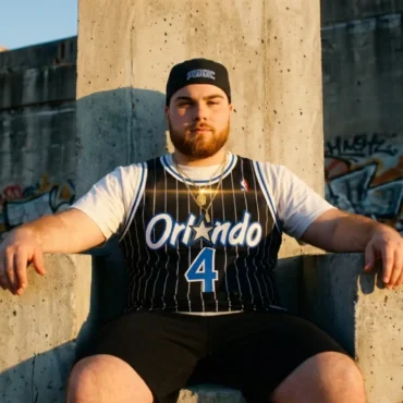 KJ Velo sitting on a concrete throne wearing an Orlando Magic jersey in an urban setting.