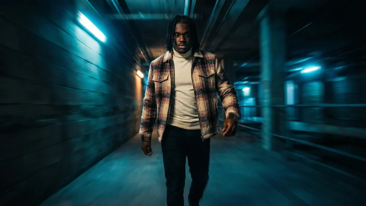 A man with dreadlocks wearing a flannel jacket walks down a dark hallway with blue lights.