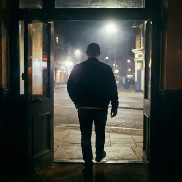 A man in a jacket and cap walking out of a dark pub into a misty street at night.