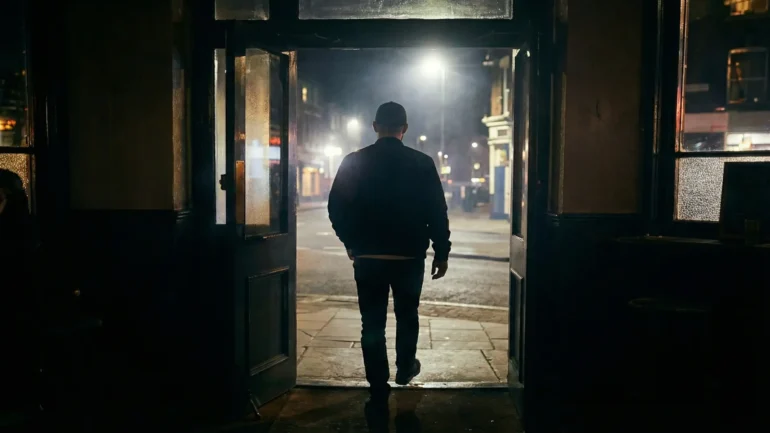 A man in a jacket and cap walking out of a dark pub into a misty street at night.