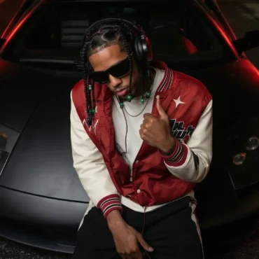 Artist Baby Draco leaning on a matte black Lamborghini Murciélago.