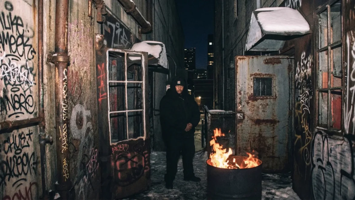 Rapper in black hoodie in snowy graffiti alley with fire barrel.