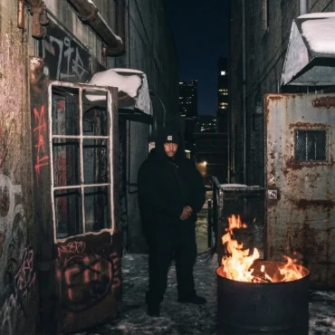 Rapper in black hoodie in snowy graffiti alley with fire barrel.