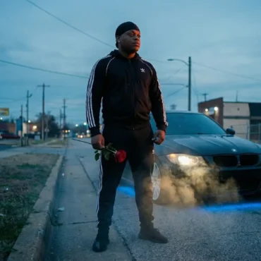 Rese2Real standing in the street holding a red rose next to a car.