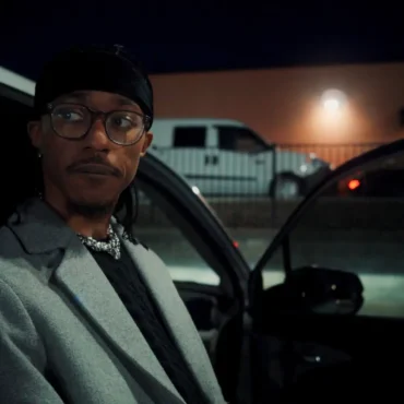 Artist JHN JSPH in a gray coat and glasses standing by a car at night.
