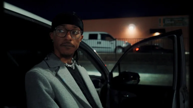 Artist JHN JSPH in a gray coat and glasses standing by a car at night.