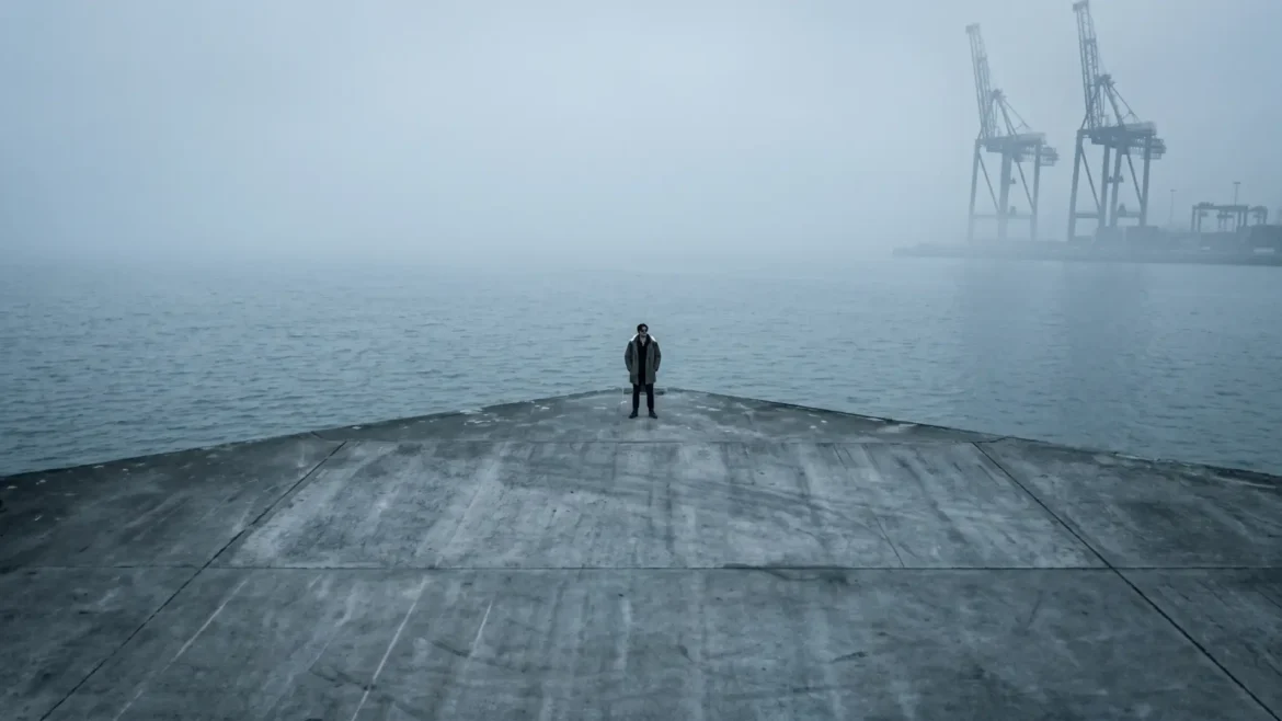 Long shot of artist B-Rain standing alone and centered at the tip of a large concrete pier in heavy fog. Behind him is a foggy ocean and part of a container port.