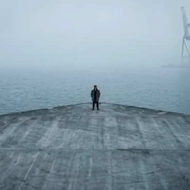 Long shot of artist B-Rain standing alone and centered at the tip of a large concrete pier in heavy fog. Behind him is a foggy ocean and part of a container port.
