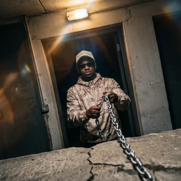 Hip hop artist Be King holding a metal chain in a dim concrete room.