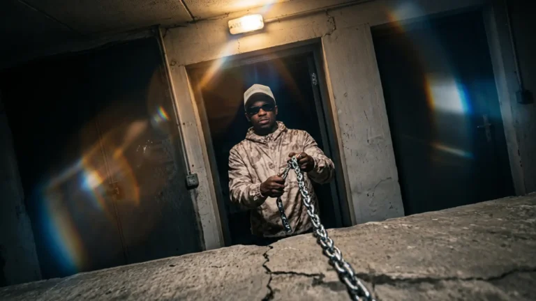 Hip hop artist Be King holding a metal chain in a dim concrete room.