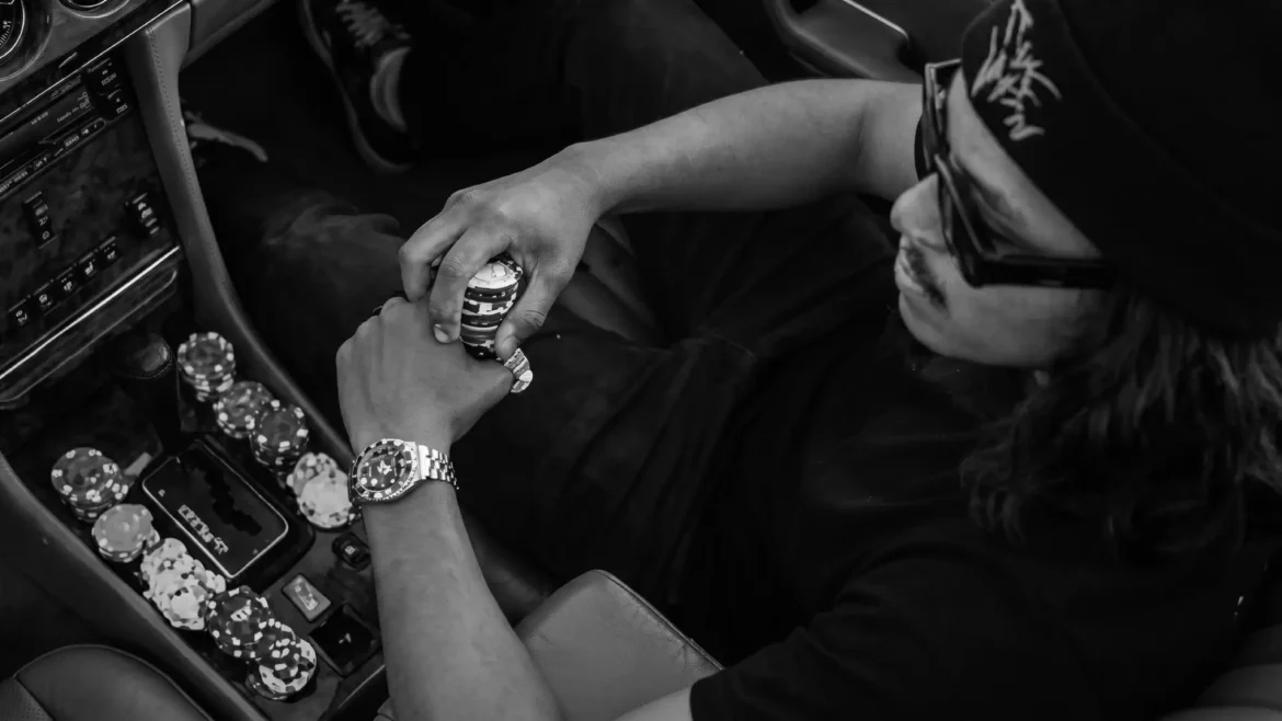 Top-down view of a man wearing a black beanie and sunglasses sitting in a brown leather car interior, holding casino poker chips near the gear shift.